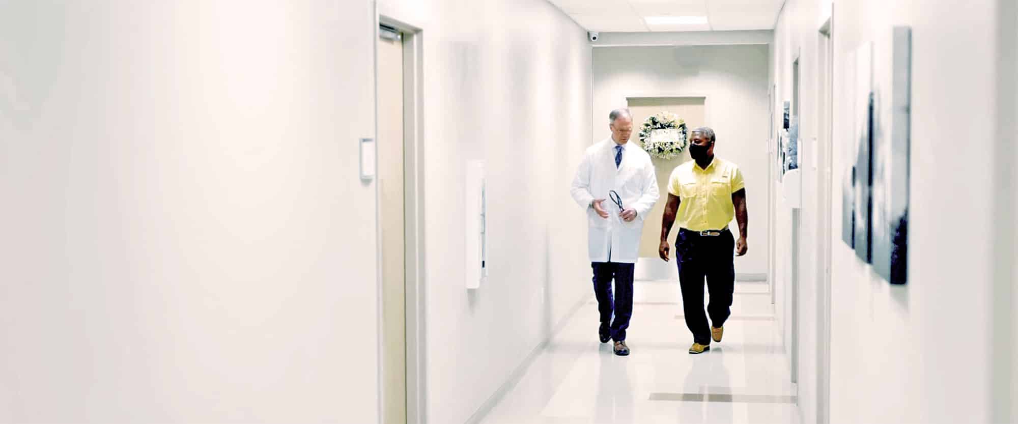 A physician and a man wearing a face mask walk down a hallway in a doctor’s office while conversing